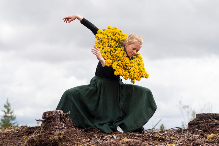 Image from the branch, the fork, the harrow on Galway Arts Centre website. Image of dancer with blonde hair wearing a long green dress. Dancer is wearing a bunch of yellow flowers around their shoulders.