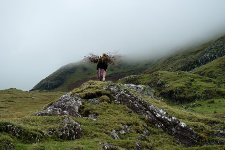 Image by Carrying Wood, on Galway Arts Centre website. Image of a person walking across green hills and land. They are carrying branches on their back.