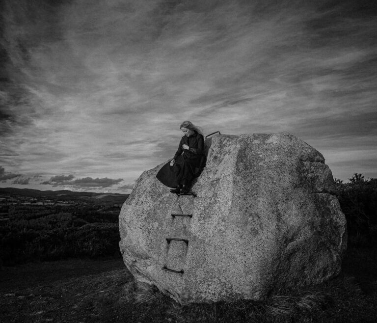 Black and white image of Ceara sitting on a large scale boulder with nature landscape in the background.