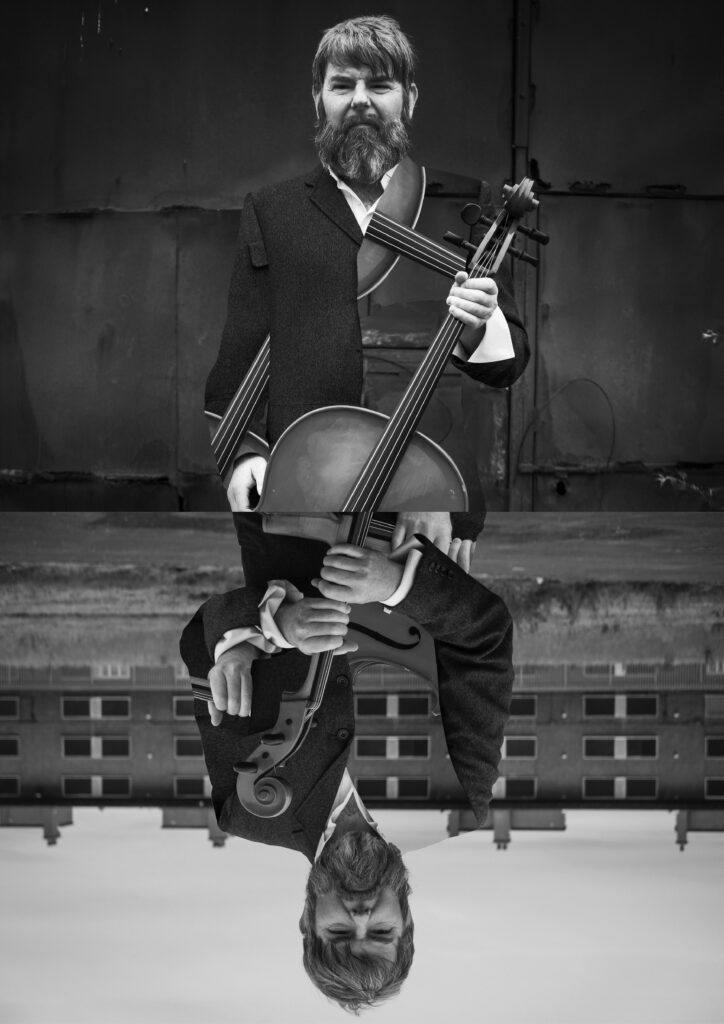 Musician Kevin Murphy standing and holding a cello. Black and white image.