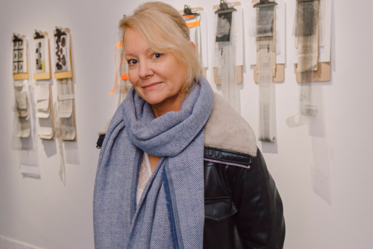 Image of Martina O'Brien, a visual artist with shoulder-length blonde hair and fair skin. She is standing in front her work at draft fissure exhibition at Galway Arts Centre.